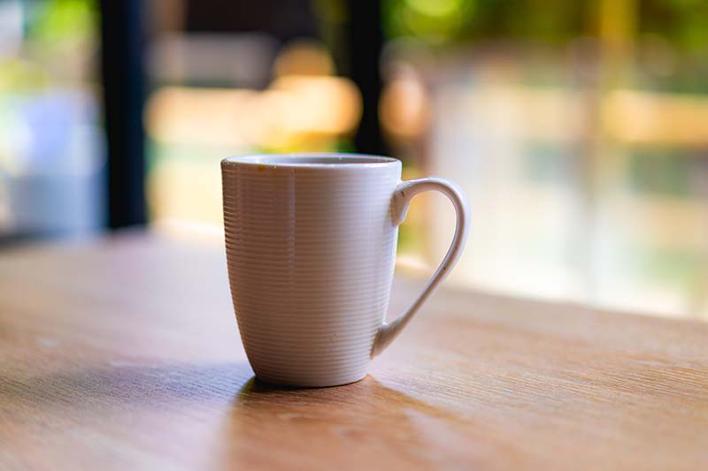 White coffee or tea mug on a wooden desk next to a window with trees in the background