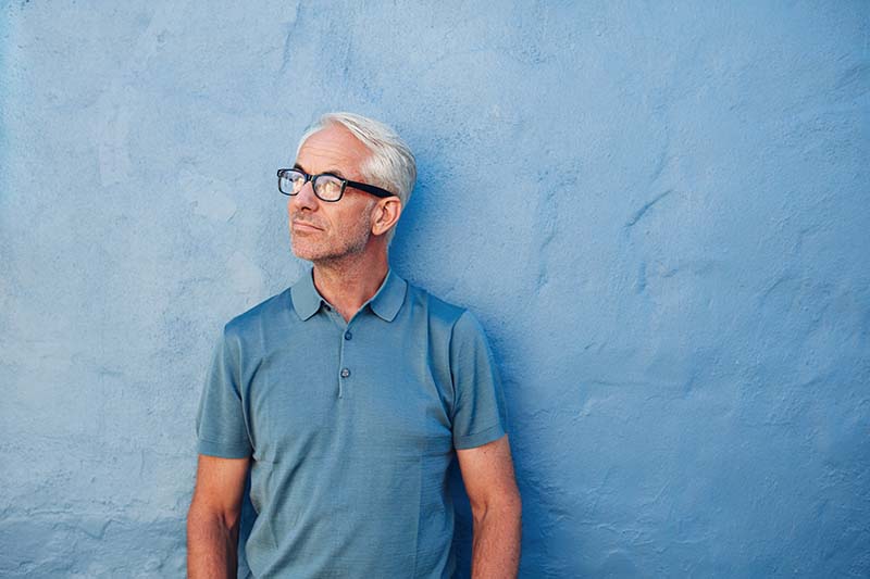 Portrait of a pensive mature man standing against a blue wall and looking away. Caucasian mid adult male with glasses.
