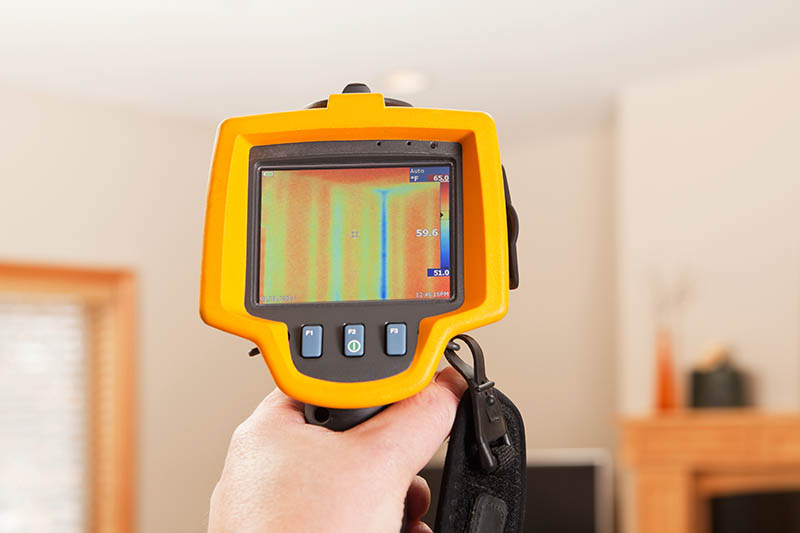 An infrared thermal imaging system being used during a home energy audit. The camera is pointed to a house living room wall and shows a distinct blue (cold) area within the home’s insulation. The center target area reads 59.6 degrees with a range of 51 to 65 degrees in the area. The blue area is a stud behind the wall, this is a typical reading in any stick built home. The wood's R-value is less than the insulation so the camera reads cooler in this area. Energy audits are performed to determine how efficient the house is and to suggest steps to increase energy efficiency. To the left is a window frame, the camera is pointed above a TV with a fireplace mantle on the right.