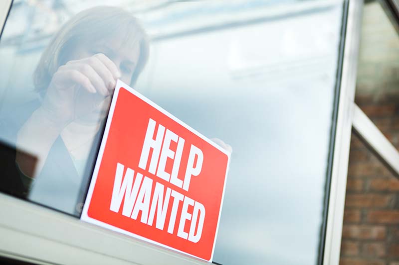 Sign of hiring, recruitment, employment and economic recovery through a woman business person posting a "HELP WANTED" sign for new employees.