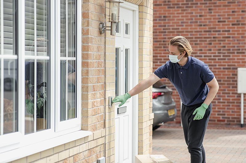 A shot of a delivery man arriving at a residential home in Northeastern, England. He is wearing protective gloves and a face mask and pressing the doorbell.