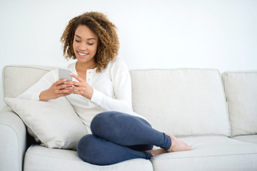 African American woman at home texting on her smart phone while lying on the couch