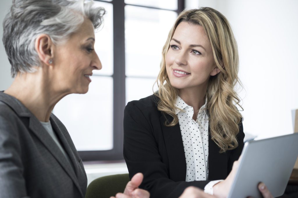 Attractive mid adult businesswoman with long blonde hair talking to mature colleague with short grey hair in office.
