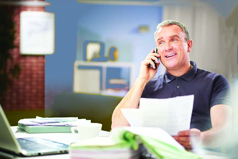 a mature man sits at his dining table at home looking through some bills and paperwork , whilst chatting happily on the phone . He is either running his business from home or just doing his home accounts. He is laughing with the customer services representative on the phone
