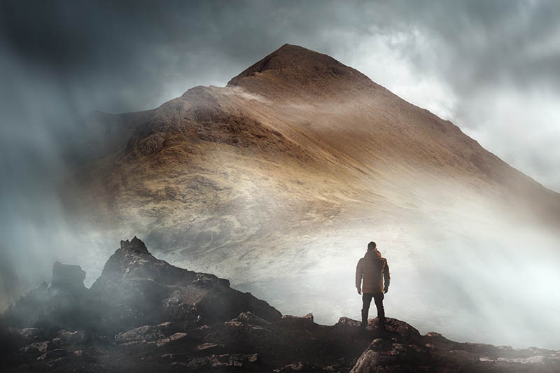 A person hiking looks onwards at a mountain shrouded in mist and clouds with the peak visible. Scenic landscape photo composite.