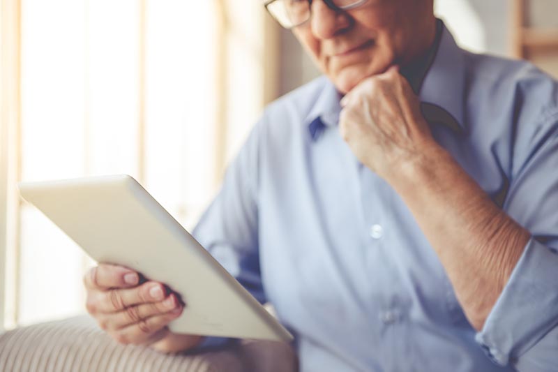 Cropped image of handsome old man in eyeglasses using a digital tablet while sitting on couch at home