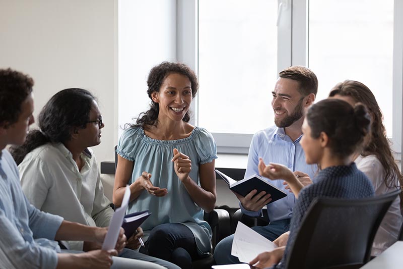 Happy diverse addicts sitting on chairs in circle, talking Happy diverse addicts sitting on chairs in circle, talking on group therapy meeting, discussing addiction, mental health problems. Multiethnic employees brainstorming on team training