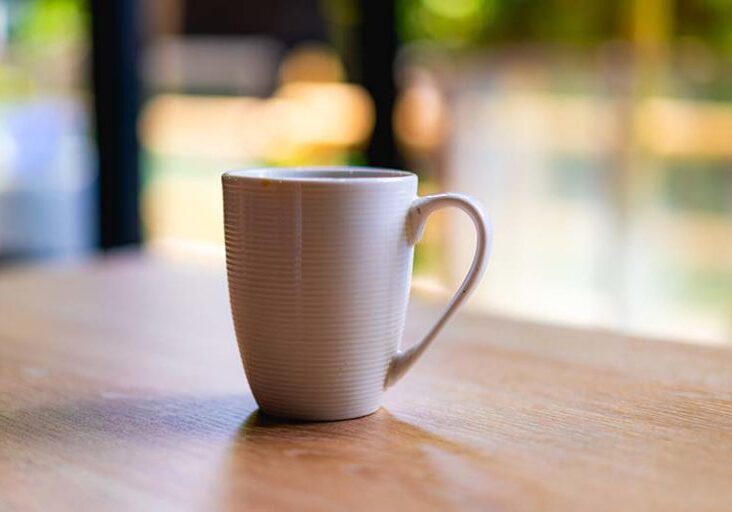 White coffee or tea mug on a wooden desk next to a window with trees in the background