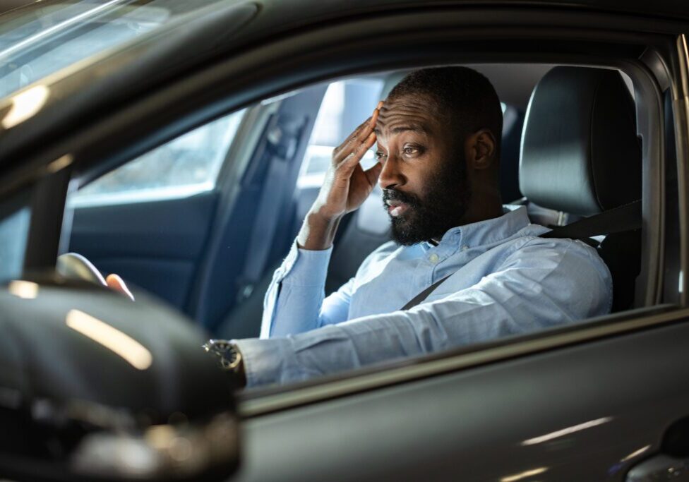 A man sits in his car at night with a stressed and concerned expression, holding his hand to his forehead. The scene conveys emotions of worry, tension, or frustration experienced in a personal moment.
