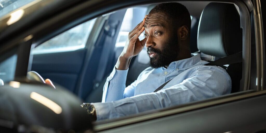 A man sits in his car at night with a stressed and concerned expression, holding his hand to his forehead. The scene conveys emotions of worry, tension, or frustration experienced in a personal moment.