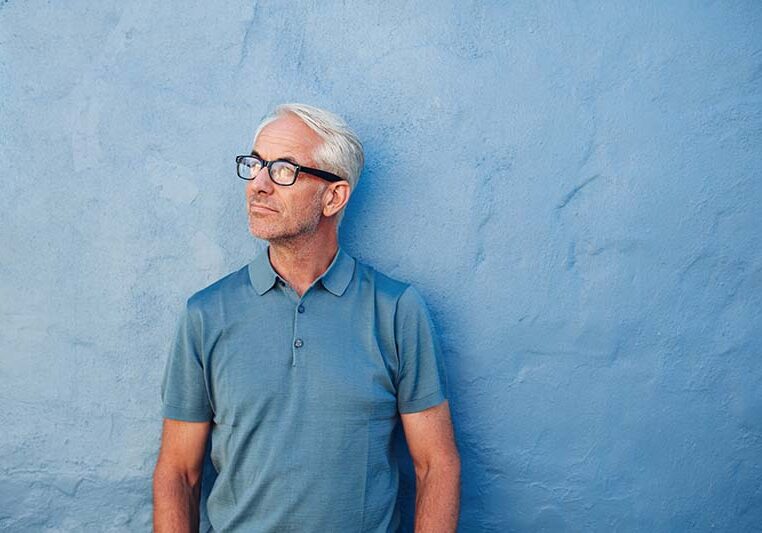 Portrait of a pensive mature man standing against a blue wall and looking away. Caucasian mid adult male with glasses.