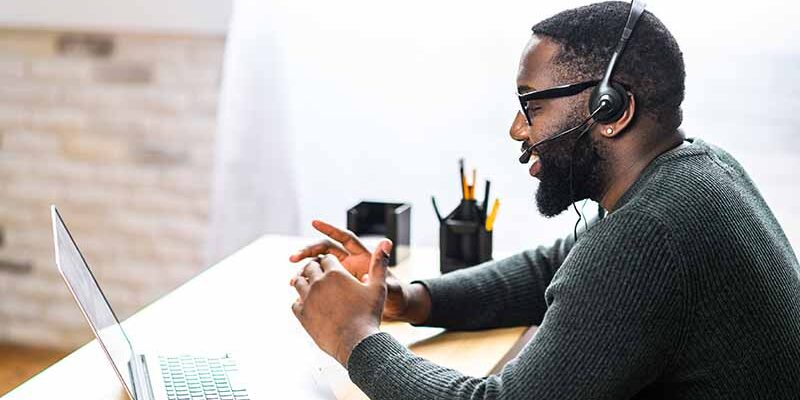 Concentrated young guy in smart casual wear and stylish glasses is using headset and laptop for online communication, supporting, selling. A black guy sits at the office desk, side view