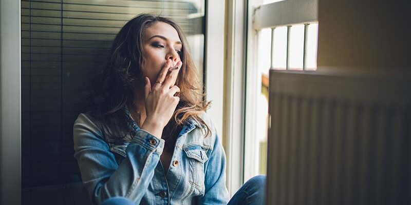 Pensive young girl sitting next to the open door and smoking a cigarette.