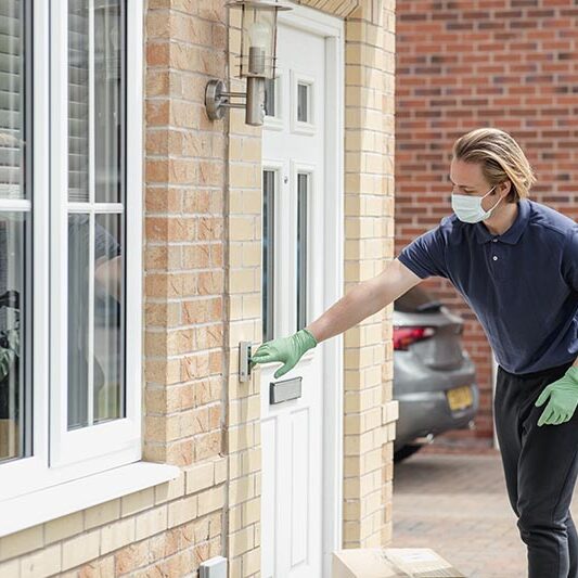 A shot of a delivery man arriving at a residential home in Northeastern, England. He is wearing protective gloves and a face mask and pressing the doorbell.