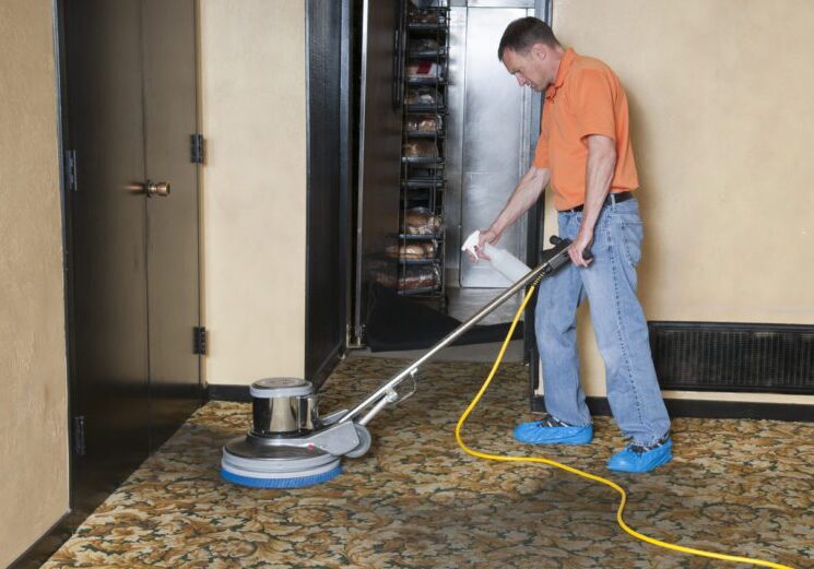 Man using a professional carpet scrubber on dirty walkway area leading from a commercial kitchen into a banquet room.