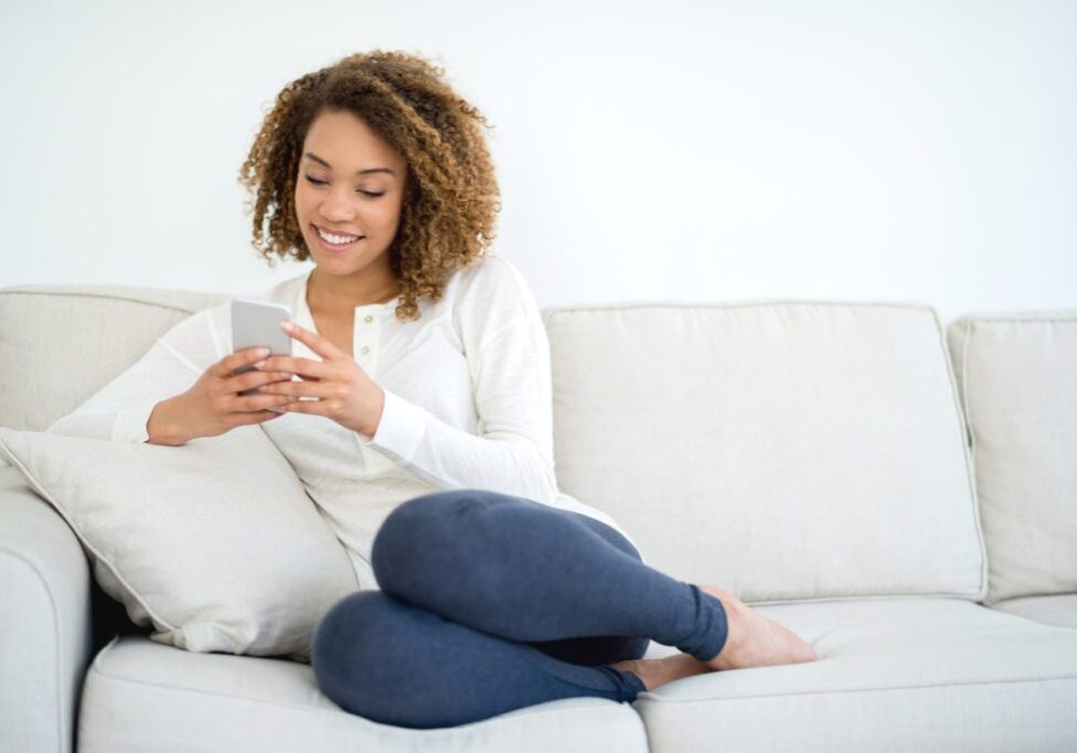 African American woman at home texting on her smart phone while lying on the couch