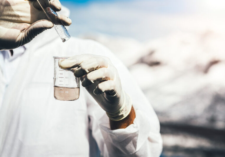 Environmental inspector in white safety suit examing polluted water at industrial site. Heaps of coal covered with white ash in the background.