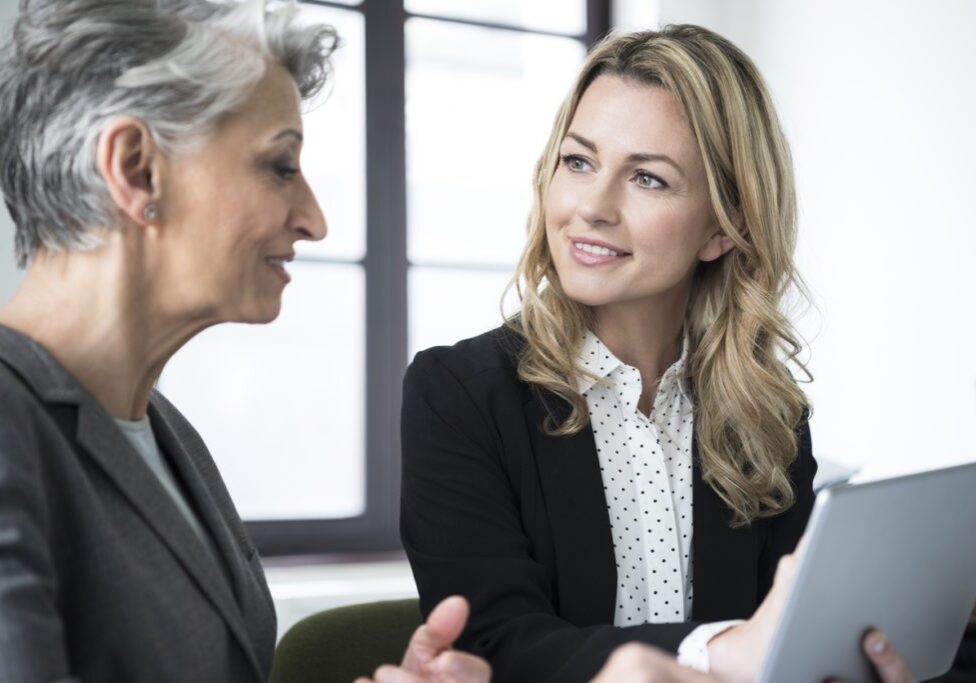 Attractive mid adult businesswoman with long blonde hair talking to mature colleague with short grey hair in office.