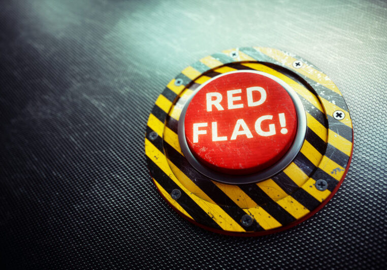 A 3D render of a conceptual mechanism with a red button and the words "RED FLAG" written on it, placed on an dark industry grid. The image can represent various concepts of danger, from political, military.. to dangers in realtionships - the so called "red flag" metaphor.