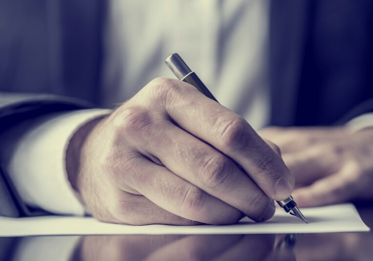 Man signing a document or writing correspondence with a close up view of his hand with the pen and sheet of notepaper on a desk top. With retro filter effect.