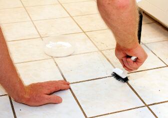 Two caucasian male hands cleaning kitchen grout of an old, dirty tile floor with environmentally friendly hydrogen peroxide, baking soda and a scrub brush.