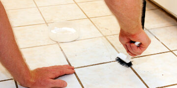 Two caucasian male hands cleaning kitchen grout of an old, dirty tile floor with environmentally friendly hydrogen peroxide, baking soda and a scrub brush.