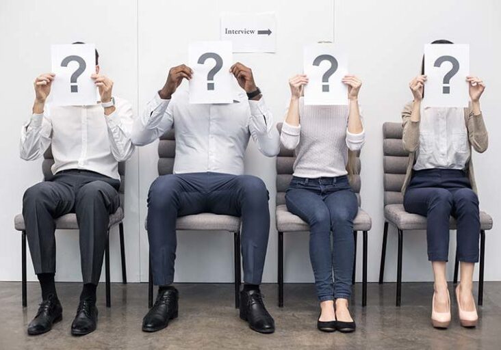 Time for job interview. Young men and women in office. They sitting, holding papers with question sign in front of faces and waiting for job interview. Nice light interior