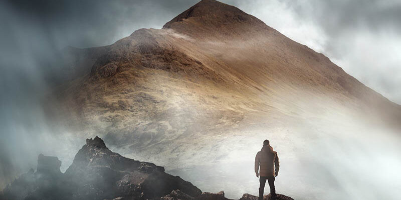 Man Hiking On A Misty Mountain A person hiking looks onwards at a mountain shrouded in mist and clouds with the peak visible. Scenic landscape photo composite.
