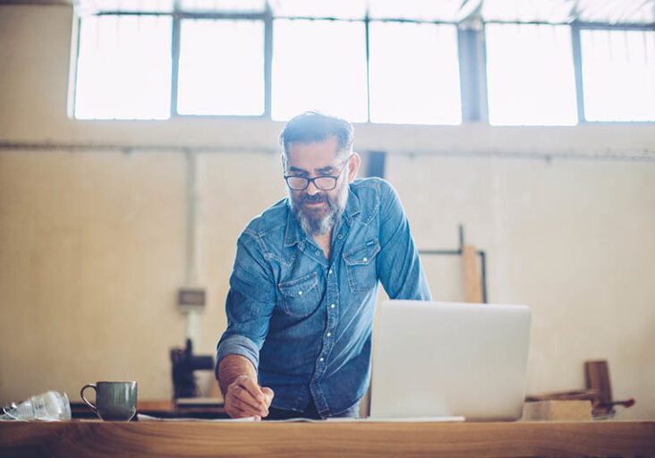 Carpenter in wood workshop using laptop and working on project. Mature man i casual clothing. Space is full of working tools and wooden planks.